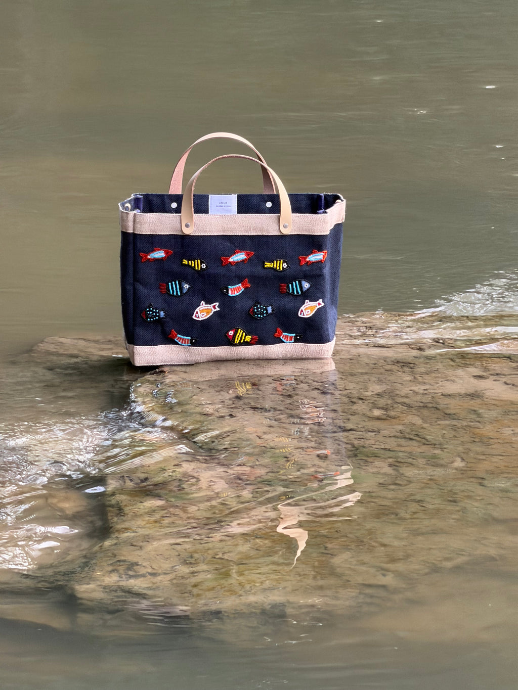 Navy Petite Market Bag with multicolored beaded fish, cream trim, and light leather handles, photographed beside a creek.