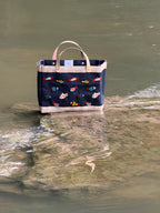 Navy Petite Market Bag with multicolored beaded fish, cream trim, and light leather handles, photographed beside a creek.